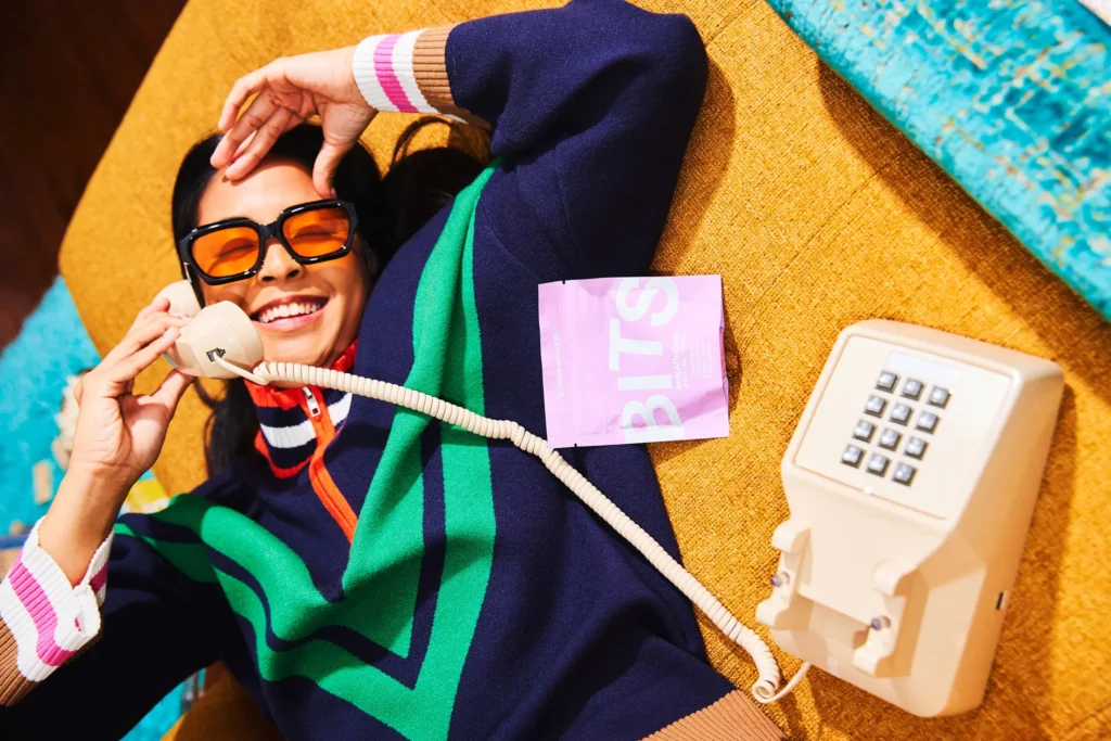 Cannabis and Productivity: Finding the Right Balance 5 Person lounging on a mustard couch in a colorful sweater, talking on a vintage corded phone with a pink BITS edible package nearby.